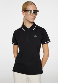 Svart pikétröja med dragkedja i halsen, vit krage och ärmdekaler, tillverkad av lättviktig tyg. Modellen bär stora solglasögon.