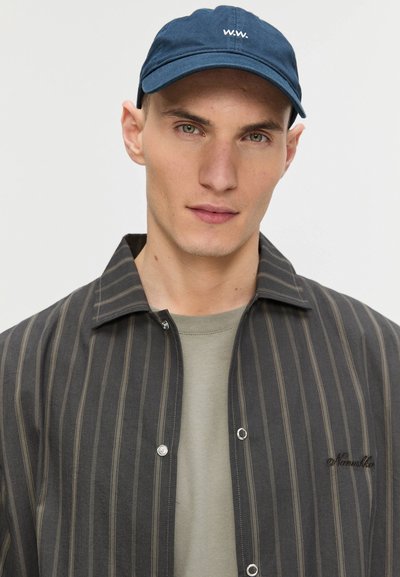 Chemise à boutons rayée noire et grise avec un t-shirt gris en dessous. Casquette bleu marine avec logo blanc. Texture fine, design à coupe normale.