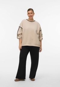 Beige sweatshirt met zwarte bies, oversized pasvorm en geribbelde manchetten, gedragen met zwarte wijdvallende broek. Model draagt bruin schoeisel.