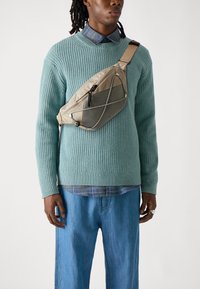 Beige nylon crossbody bag with mesh panel, adjustable strap, and zipper closure. Worn over a teal knitted sweater and denim pants.