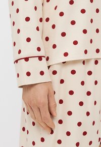 Béžová pyžamová sada s červenými puntíky, která obsahuje dlouhý rukáv top a sukni s elastickým pasem. Hladká bavlněná textura a kontrastní lemování.