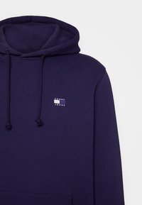 Hanorac bleumarin cu glugă ajustabilă, având un mic patch logo Tommy Jeans pe piept și un buzunar de tip kangaroo în față.