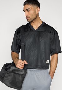 Μαύρο αθλητικό μπλουζάκι από δίχτυ με λαιμόκοψη V και κοντά μανίκια, φορεμένο με γκρι σορτς. Κρατάει μια μαύρη τσάντα duffle με μπροστινή τσέπη.