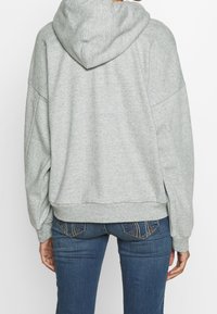 Person wearing a light gray hooded sweatshirt and blue jeans, shown from the back against a plain white background.