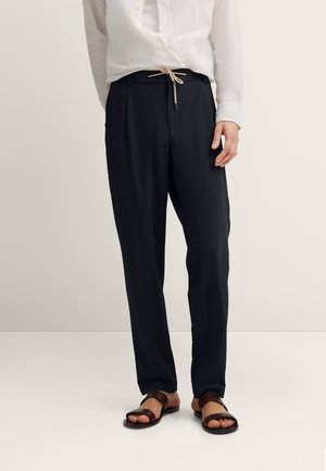 Mann trägt schwarze, schmal zulaufende Hose mit beiger Schnur, weißes Hemd mit Knöpfen und dunkelbraune Ledersandalen, steht vor neutralem Hintergrund.