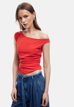 Kvinde iført en rød ensiddet top med rynker og løstsiddende blå jeans med bælte, stående mod en ensfarvet baggrund.
