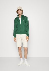 Veste zippée verte avec des rayures blanches sur les côtés, associée à un short beige clair. Le modèle porte une casquette beige et des sneakers blanches.
