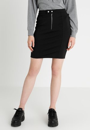 Jupe crayon noire avec une fermeture éclair à l'avant et deux boutons-pression à la taille, fabriquée dans un tissu extensible. Portée avec un haut gris et des baskets noires.
