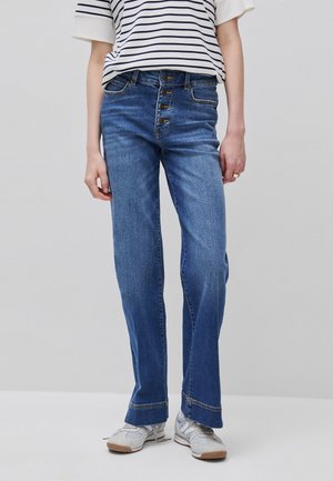 Persona che indossa jeans blu a vita alta con quattro bottoni visibili, abbinati a una maglietta a righe e sneakers bianche su uno sfondo semplice.