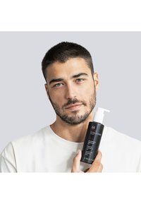 Czarna butelka żelu oczyszczającego z białą pompką, opisana czerwonym i białym tekstem. Modelka w białej koszuli trzyma produkt, pokazując jego szczegóły.