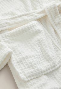 Balta, strukturēta peldmētelis ar vafeļu rakstu, ar mīkstu, pūkainu sajūtu un ērtu kabatu. Liesmas dizains ar jostu.