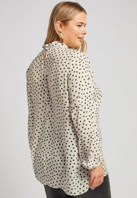 Blusa beige con pois neri, maniche lunghe e dettaglio del collo arricciato. Tessuto morbido con una vestibilità leggermente ampia e orlo arrotondato.