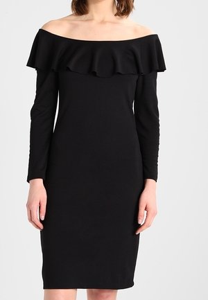 Vrouw die een zwarte off-shoulder jurk tot op de knie draagt met lange mouwen en een gerimpelde halslijn tegen een effen achtergrond.