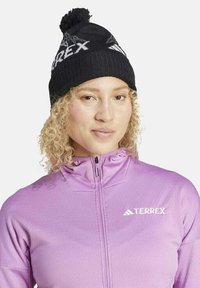 Donna con capelli biondi ricci che indossa un berretto nero con pompon e una giacca Terrex viola con cerniera, vista frontalmente con un'espressione neutra.