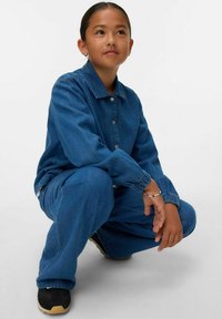 Denim obleka v temno modri barvi, ki vključuje jakno s gumbki in hlače sproščenega kroja. Gladka tekstura z elastičnimi manšetami in črnimi čevlji.
