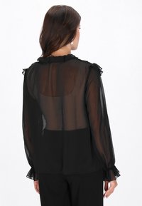 Fekete áttetsző blúz fodros gallérral és mandzsettákkal, hosszú ujjakkal és laza szabással, enyhén átlátszó hátrészlettel.