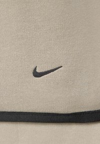 Beige-kangas, jossa on teksturoitu pinta, sisältää mustan brodeeratun Nike-swoosh-logon, joka on sijoitettu keskelle, ja jota korostaa musta vaakaraita lähellä alaosaa.