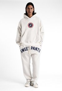 Ensemble de sweat à capuche et de joggings en polaire beige, avec impression "SWEET PANTS", patch rond et accents contrastants bleu foncé.