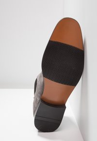 Chaussure en suède gris avec une semelle en caoutchouc noire. La semelle présente un motif texturé et une section marron contrastante au niveau du talon.