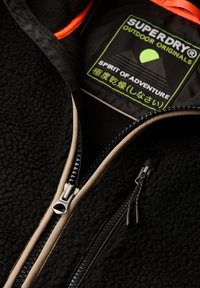 Czarna polarowa kurtka z wysokim kołnierzem, kontrastowym beżowym zamkiem błyskawicznym, metalowym zamkiem i zieloną metką z tekstem oraz grafikami gór.