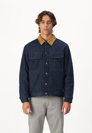 Giacca di jeans blu scuro con colletto in corduroy beige, dotata di due tasche sul petto e bottoni argentati, abbinata a pantaloni grigi chiari.