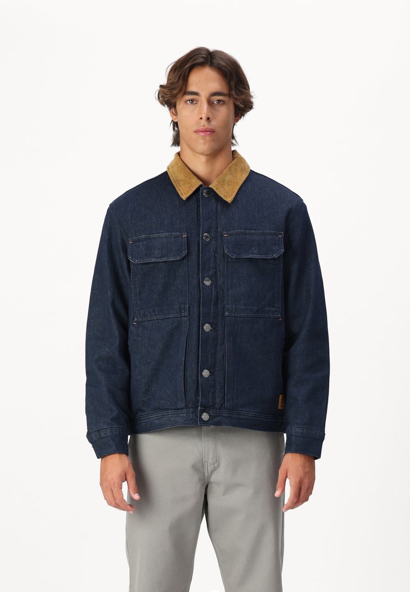 Jeansowa kurtka w ciemnoniebieskim kolorze z beżowym kołnierzem z korduroy, z dwoma kieszeniami na piersi i srebrnymi guzikami, w połączeniu z jasnoszarymi spodniami.