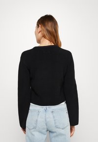 Schwarzer Strickpullover mit kurzer Passform, gerippter Textur, rundem Ausschnitt und langen Ärmeln, getragen mit hellblauen Jeans, Rückansicht.