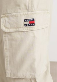 Vaaleanbeige cargo-housut, joissa on sivutasku Tommy Jeans -logolla, ommeltuja yksityiskohtia ja sileä, kevyt rakenne.