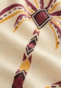 Tissu beige avec un motif floral symétrique imprimé en rouge foncé, jaune moutarde et formes noires.
