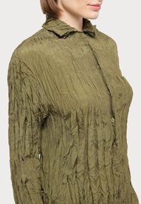 Olive green, crinkled button-up shirt with a pointed collar and vertical texture. Features seven front buttons and a slim fit design.