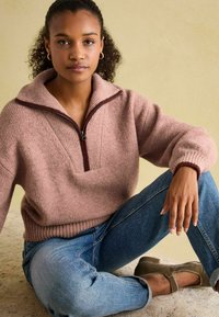 Chunky, hellrosa Strickpullover mit einem Halbreißverschluss-Kragen und gerippten Bündchen, kombiniert mit blauen Jeans und taupefarbenen Schuhen vor einem neutralen Hintergrund.