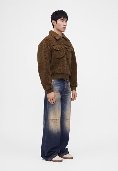 Homme portant une veste marron duveteuse et un jean bleu délavé à jambes larges, debout sur fond blanc.