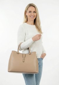 Blonde Frau trägt weißen Pullover und blaue Jeans, hält eine große beige Einkaufstasche mit goldener Schnalle, lächelt vor weißem Hintergrund.