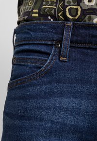 Mörkblå denimjeans med klassisk femfickedesign, kontrasterande orange stickning och en slät, medeltjock textur.