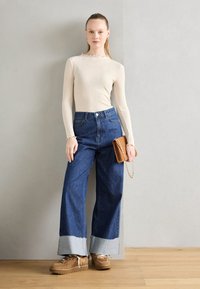 Beige geripptes Langarmoberteil mit gewellten Kanten, kombiniert mit hochgeschnittenen dunkelblauen Schlagjeans mit hellblauem Saum. Braune Plateauschuhe und eine sandfarbene Clutch.