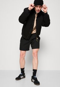 Black bomber jacket with a zip, beige sweatshirt underneath, black shorts, and black sneakers with white stripes; black beanie completes the look.