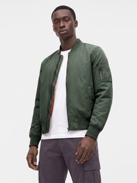 Jeune homme portant une veste bomber verte sur un t-shirt blanc et un pantalon cargo foncé sur un fond clair uni.