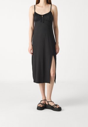 Robe noire sans manches avec une encolure froncée, détail en forme de trou et fente sur le côté. Texture lisse, coupe ajustée, assortie à des sandales noires à lanières.