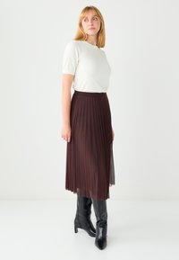 Jupe midi plissée marron avec une taille lisse, associée à un haut crème clair à manches courtes et des bottes à talons noires montant jusqu'aux genoux.