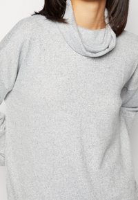 Maglione grigio chiaro con collo a cowl realizzato in un morbido tessuto texturizzato, caratterizzato da maniche lunghe e una vestibilità rilassata. I dettagli includono un sottile motivo a maglia.