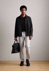 Man with afro hairstyle walking, wearing black jacket, black shirt, light pants, black shoes, and carrying a black leather bag.