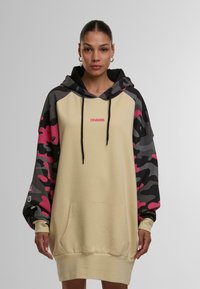 Bež hoodica oversize z rokavi v črno-roza kamuflaži, s kapuco na vrvico, sprednjim žepom in roza logotipom na prsih. Material iz mešanice bombaža.