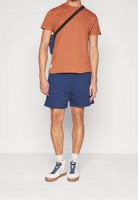 T-shirt orange à col rond, shorts bleu marine, baskets montantes blanches avec accents bleu marine, et un petit sac en bandoulière bleu. Tenue décontractée pour temps chaud.