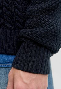 Suéter de punto azul oscuro con patrones texturizados y puños acanalados, resaltando una textura gruesa y acogedora, además de elementos de diseño robustos.