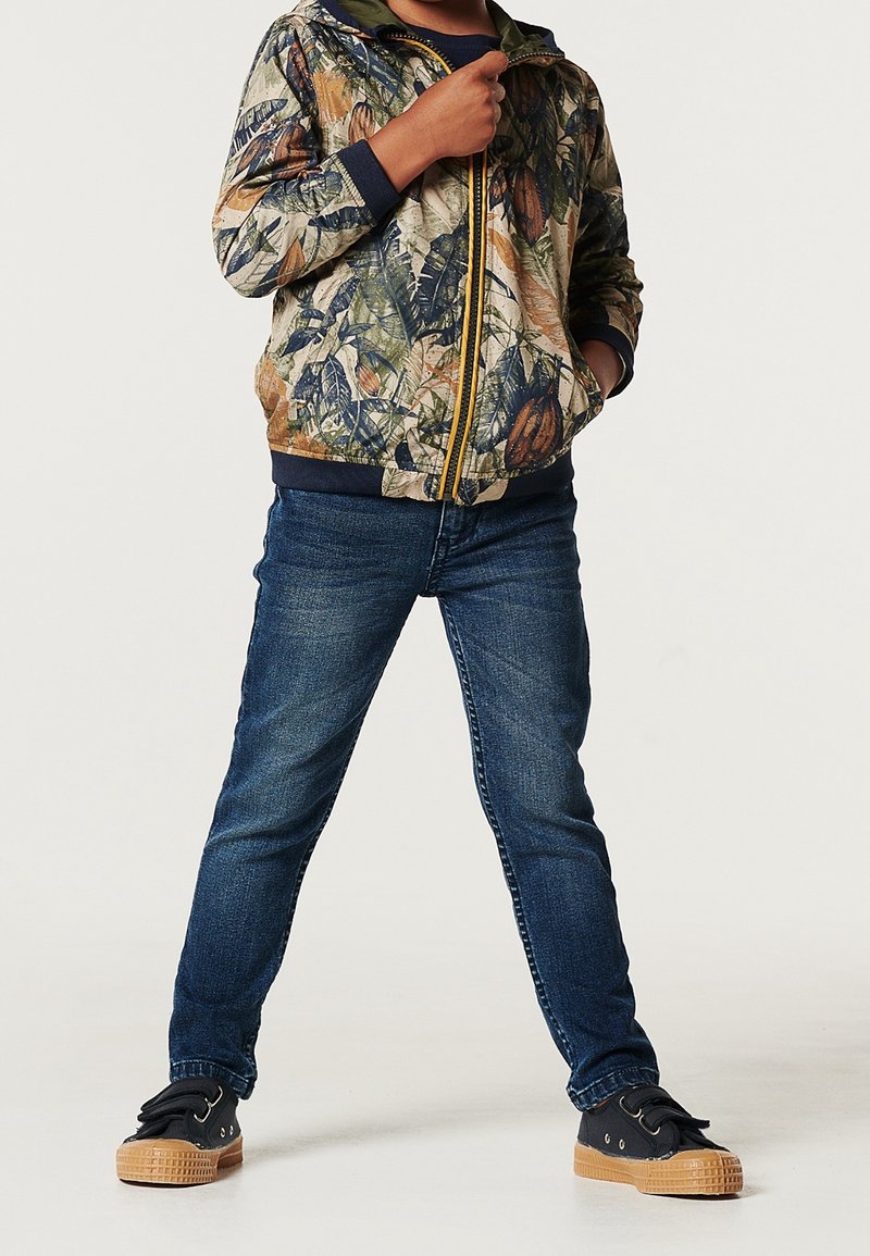 Camo-mönstrad zip hoodie i gröna och jordnära nyanser, kombinerad med mörkblå jeans och svarta sneakers med beige sulor.