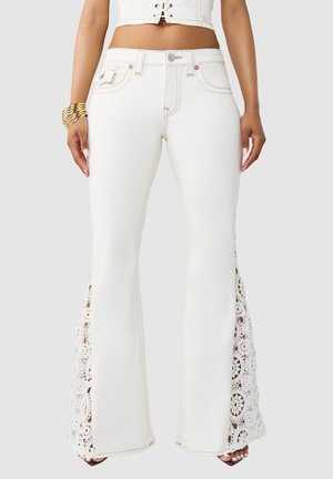 Vrouw draagt witte uitlopende jeans met kanten haakwerkpanelen aan de buitenkant van de benen, gecombineerd met gouden armbanden en een wit cropped topje.