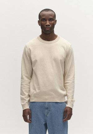 Mann trägt einen cremefarbenen Pullover mit langen Ärmeln und weiten blauen Jeans, steht vor einem schlichten weißen Hintergrund und blickt geradeaus.