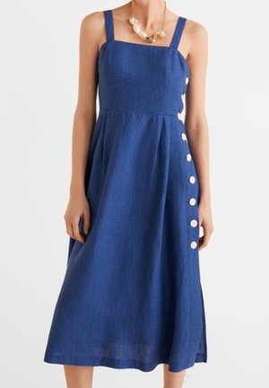 Mouwloze blauwe midi-jurk met brede bandjes, vierkante halslijn, zijknopen en geplooide rok, gedragen met een schelpenketting.