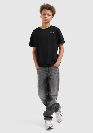 Tienerjongen met krullend haar, gekleed in een zwart T-shirt, grijs vervaagde spijkerbroek en witte sneakers, staat met zijn handen in zijn zakken.