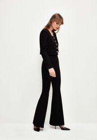 Zwarte cropped jas met gouden knopen, gecombineerd met strak zittende zwarte flared pants. Soepele textuur, op maat gemaakt ontwerp en monochroom kleurenpalet.
